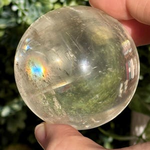 Op de afbeelding: Een heldere kwartskristallen bol, vastgehouden in een hand, met interne breuken en een regenboogeffect. De bol is rond en gepolijst en reflecteert licht en het omringende groen. Het kristal heeft een bleke, doorschijnende kleur.