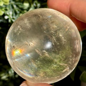 Op de afbeelding: Een heldere kristallen bol met interne insluitsels en een regenboogbreking. De bol wordt in een hand gehouden, met groene bladeren op de achtergrond. De bol heeft een diameter van ongeveer 7,6 cm.