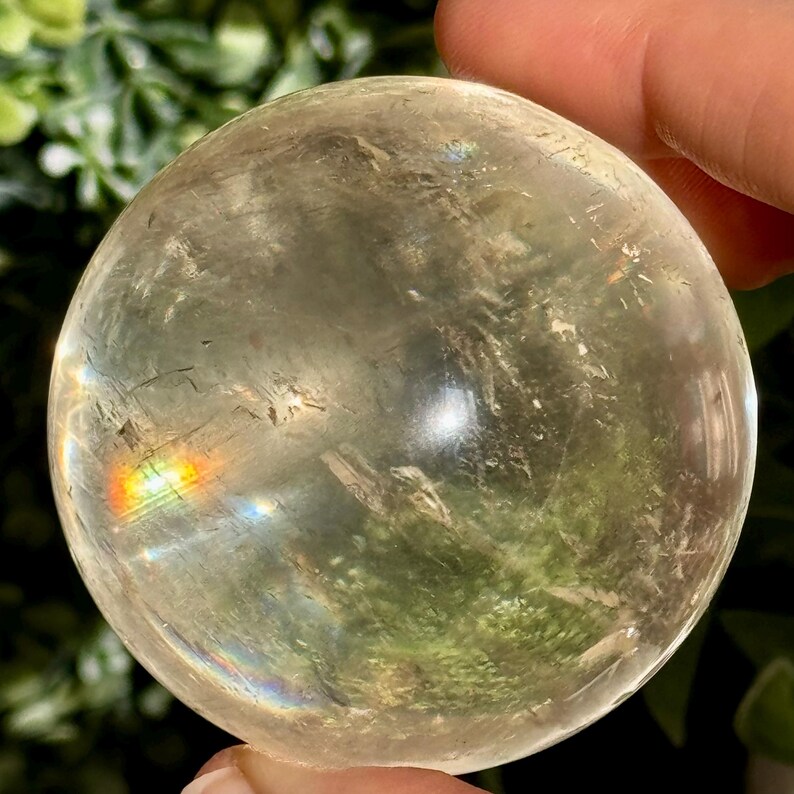 Op de afbeelding: Een heldere kristallen bol met interne insluitsels en een regenboogbreking. De bol wordt in een hand gehouden, met groene bladeren op de achtergrond. De bol heeft een diameter van ongeveer 7,6 cm.