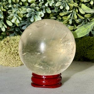 Op de afbeelding: Een heldere, bolvormige kristallen bol staat op een kleine, ronde, rode houten standaard. De kristallen bol heeft een lichtgele tint en lijkt doorschijnend. De achtergrond toont groen gebladerte en een lichtgrijze ondergrond.