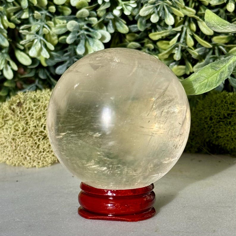 Op de afbeelding: Een heldere, bolvormige kristallen bol staat op een kleine, ronde, rode houten standaard. De kristallen bol heeft een lichtgele tint en lijkt doorschijnend. De achtergrond toont groen gebladerte en een lichtgrijze ondergrond.