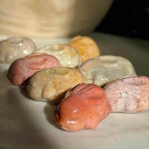 May include: A collection of carved stone bunny figurines in various colors, including coral, beige, and orange. The bunnies are small, with detailed features and a smooth, polished finish. The figurines are arranged on a white surface.