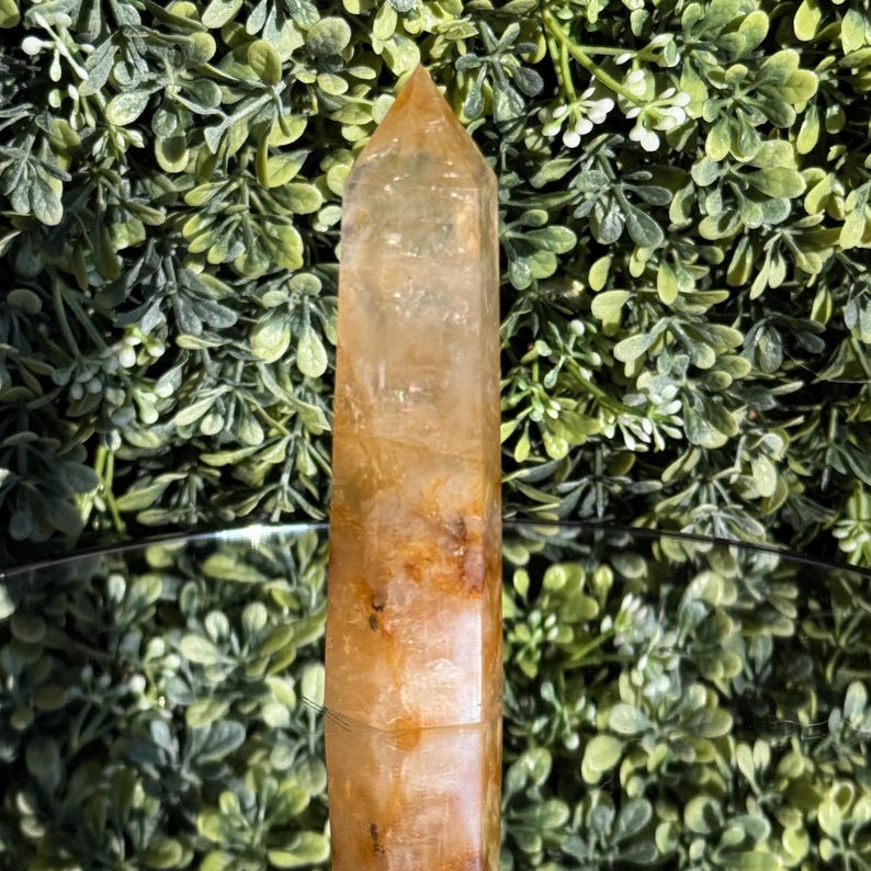 May include: A golden-yellow quartz crystal tower with a pointed top. The crystal has a rectangular shape with visible internal patterns and inclusions. The crystal is displayed against a backdrop of green foliage.