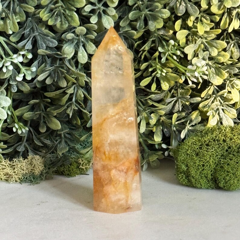 May include: A tall, polished crystal obelisk with a pointed top. The crystal is a translucent amber color with streaks of orange and white. The obelisk is standing upright against a backdrop of green foliage.