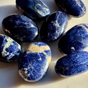 May include: A collection of eight polished sodalite stones. The stones are oval-shaped, with a deep blue color and white and tan veining. The stones are arranged on a white surface, and the lighting creates highlights and shadows on their surfaces.