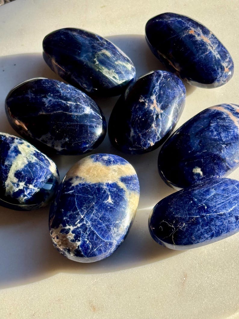May include: A collection of eight polished sodalite stones. The stones are oval-shaped, with a deep blue color and white and tan veining. The stones are arranged on a white surface, and the lighting creates highlights and shadows on their surfaces.