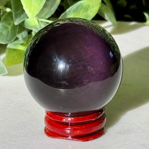 May include: A polished, dark purple sphere sits atop a vibrant red, tiered wooden stand. The sphere has a glossy finish, reflecting light. The background features green foliage, adding a natural contrast to the object.