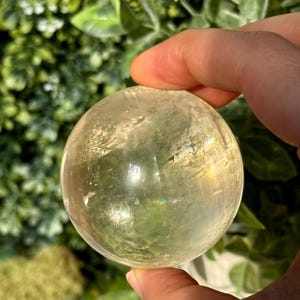 Op de afbeelding: Een heldere, geel getinte kristallen bol. Het gepolijste oppervlak reflecteert licht, waardoor een subtiel regenboogeffect ontstaat. De bol wordt in een hand gehouden, met een vage groene achtergrond van bladeren.