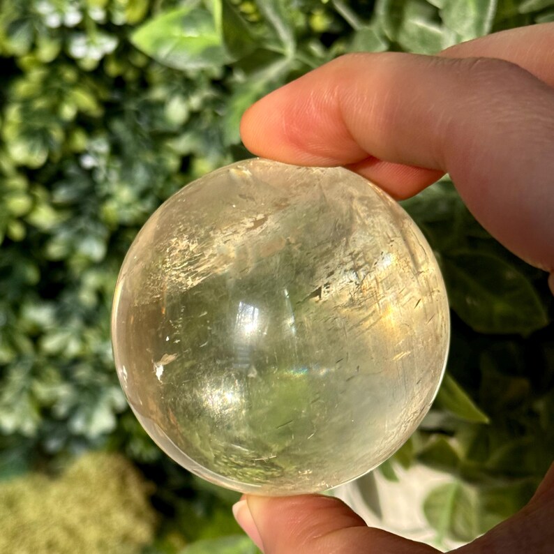 Op de afbeelding: Een heldere, geel getinte kristallen bol. Het gepolijste oppervlak reflecteert licht, waardoor een subtiel regenboogeffect ontstaat. De bol wordt in een hand gehouden, met een vage groene achtergrond van bladeren.