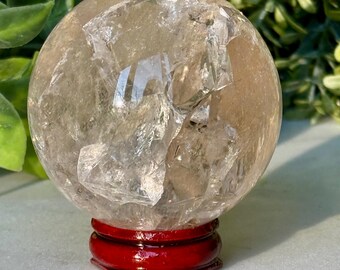 May include: A clear quartz crystal sphere with internal fractures, resting on a glossy, red wooden stand. The sphere has a translucent, light brown hue and reflects light. The stand is a deep, rich red color.