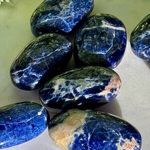 May include: A collection of polished sodalite stones. The stones are oval-shaped with a deep blue color, veined with white and hints of beige. The smooth, glossy surfaces reflect light, highlighting the natural patterns. These stones are ideal for crystal healing and meditation.