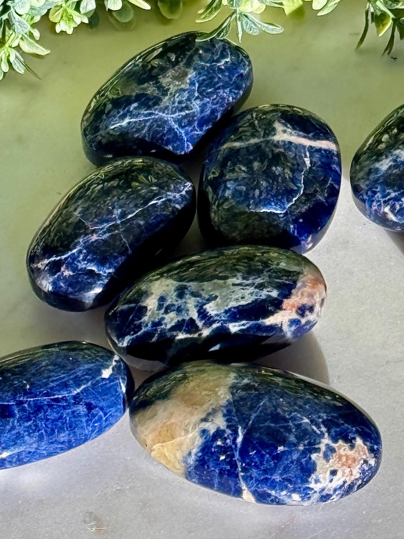 May include: A collection of polished sodalite stones. The stones are oval-shaped with a deep blue color, veined with white and hints of beige. The smooth, glossy surfaces reflect light, highlighting the natural patterns. These stones are ideal for crystal healing and meditation.