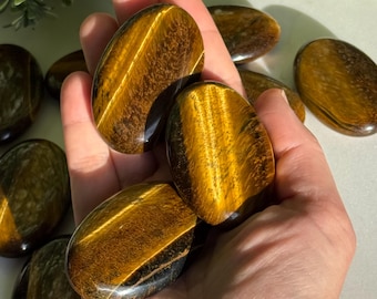 May include: A collection of polished tiger's eye stones, oval-shaped with a chatoyant effect. The stones display bands of golden brown and black, with a smooth, glossy finish. Some stones are held in a hand, while others are scattered around.