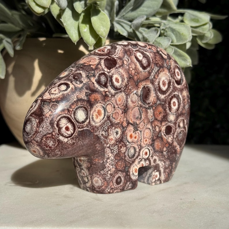 May include: A polished stone carving of a bear, crafted from orbicular jasper. The surface features a dense pattern of circular, eye-like inclusions in shades of deep brown, reddish-maroon, cream, and tan, creating a textured, mottled appearance.