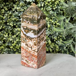 May include: A multi-colored stone obelisk with a flat base and a pointed top. The stone features layers of white, brown, orange, and green hues. The obelisk is standing upright against a backdrop of green foliage.