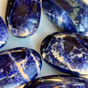 May include: A collection of polished sodalite stones. The stones are oval-shaped with a deep blue color and white and beige veining. The polished surfaces reflect light, highlighting the natural patterns and textures of the mineral.