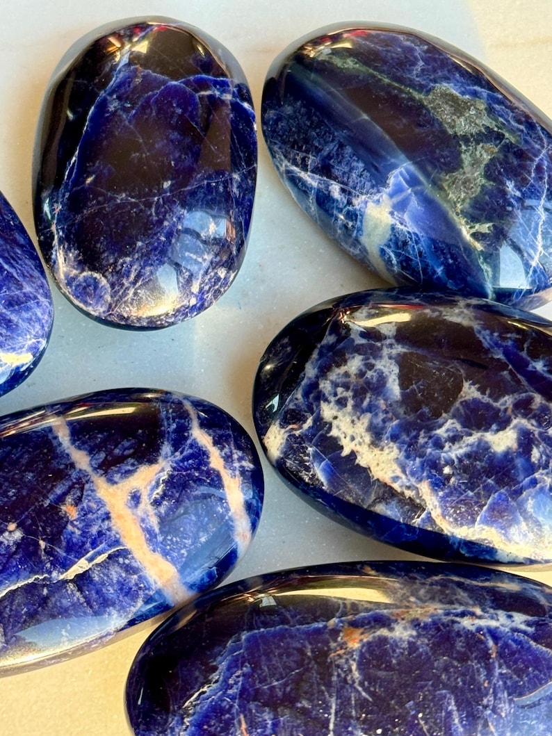May include: A collection of polished sodalite stones. The stones are oval-shaped with a deep blue color and white and beige veining. The polished surfaces reflect light, highlighting the natural patterns and textures of the mineral.