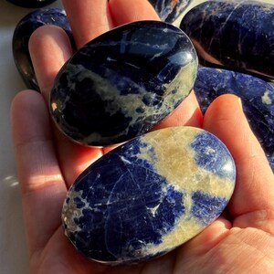 May include: Several polished sodalite stones in shades of deep blue and cream are held in a hand. The stones have a smooth, glossy finish and varying patterns of color. The stones are oval-shaped and appear to be natural gemstones.