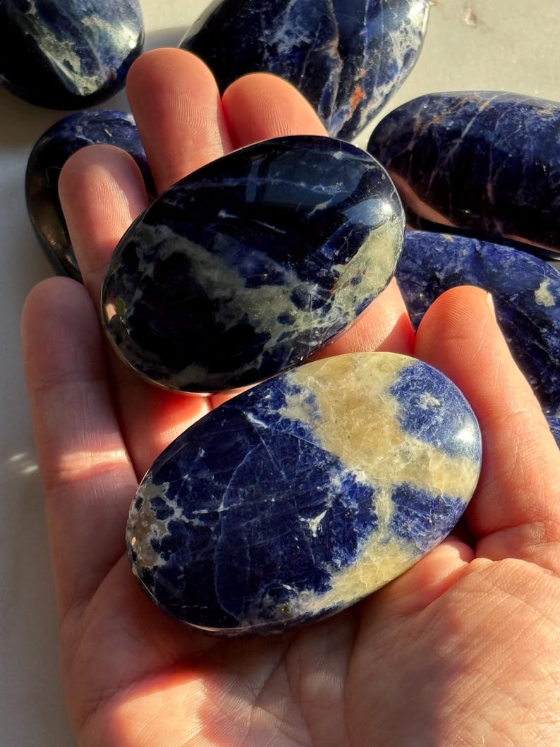 May include: Several polished sodalite stones in shades of deep blue and cream are held in a hand. The stones have a smooth, glossy finish and varying patterns of color. The stones are oval-shaped and appear to be natural gemstones.