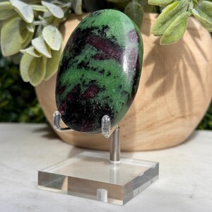 May include: An oval-shaped polished zoisite stone featuring deep green, black, and dark red mineral inclusions. The stone is displayed upright on a clear acrylic stand with two metal prongs supporting the base of the smooth, rounded mineral specimen.