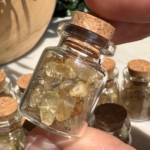May include: A small, clear glass jar with a cork stopper filled with polished, translucent yellow and golden-brown rutilated quartz crystal chips. The jar is square-shaped with rounded corners and is held up to show the contents clearly.