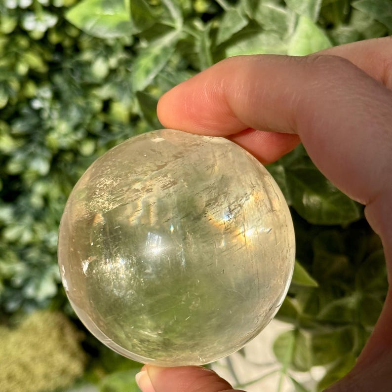 Op de afbeelding: Een heldere, bolvormige kristallen bol met een lichtgele tint. De bol wordt in een hand gehouden, met een groene, bladerrijke achtergrond. Het kristal heeft interne patronen en een lichte iriserende glans.