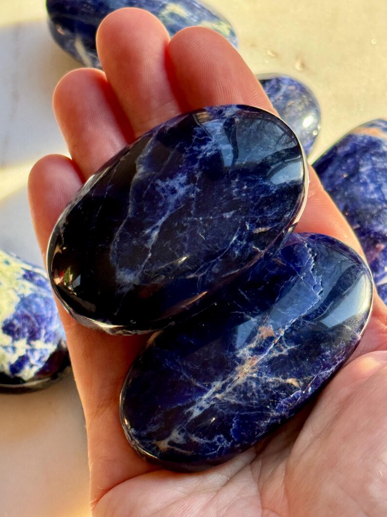 May include: Two polished sodalite stones with a deep blue color and white veining are held in a hand. Additional stones are visible in the background. The stones have an oval shape and a smooth, glossy finish.