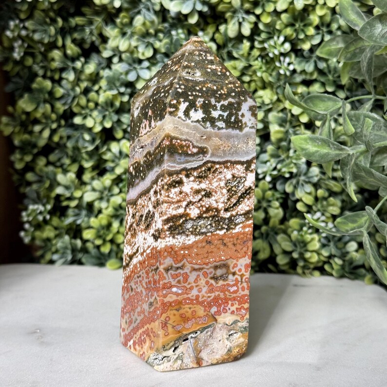 May include: A polished, rectangular gemstone obelisk with a flat base and a pointed top. The stone features bands of color, including white, brown, orange, and green, with speckled patterns. The obelisk is displayed against a backdrop of green foliage.