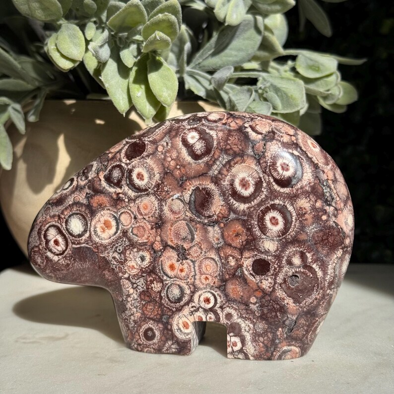 May include: A carved stone bear figurine in shades of brown, cream, and reddish-brown. The bear has a detailed pattern of circular and oval shapes. The figurine is a decorative item, possibly for a home or office.