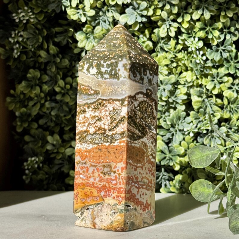 May include: A tall, rectangular crystal obelisk with a pointed top. The stone features a mix of colors, including white, green, orange, and brown, with a speckled and layered pattern. The crystal is standing upright against a backdrop of green foliage.