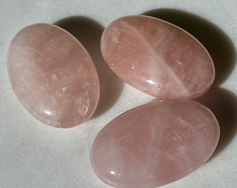May include: Three polished rose quartz stones. Each stone is oval-shaped with a smooth, glossy surface. The stones have a soft pink hue, with subtle variations in color and natural veining. The stones are arranged on a white surface.