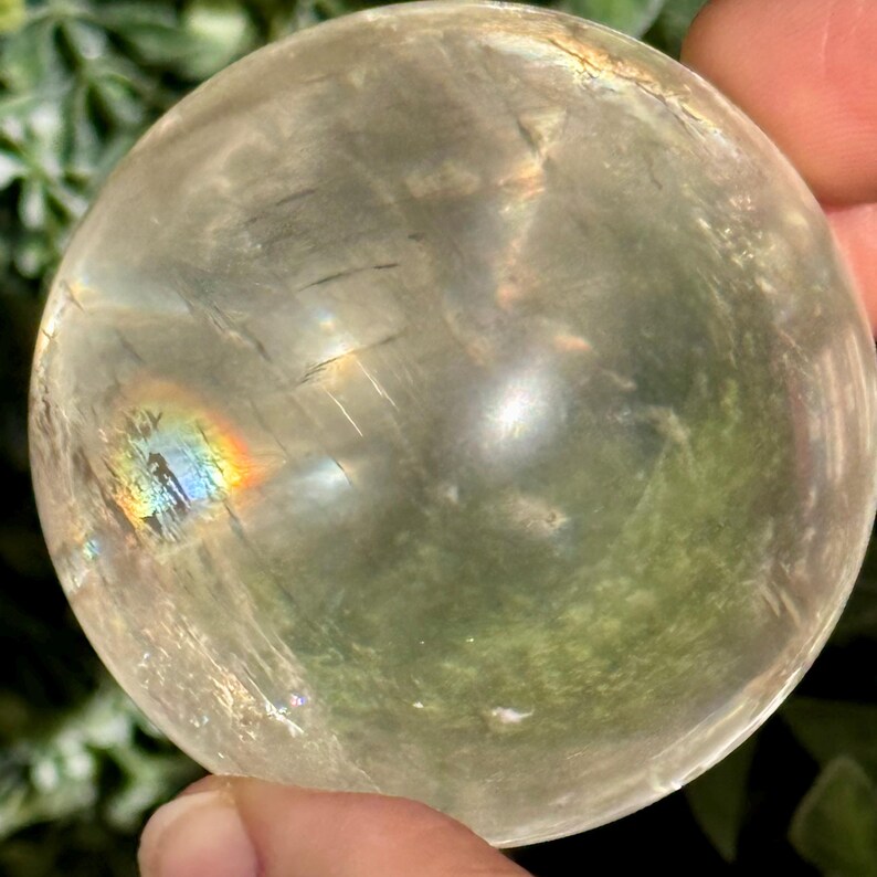 Op de afbeelding: Een heldere, bolvormige kristallen bol met interne insluitsels en een regenboogachtige iriserende glans. De bol wordt in een hand gehouden, met een vage groene achtergrond.