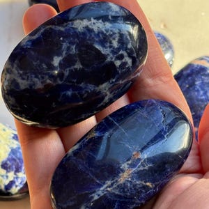 May include: Several polished sodalite stones in shades of deep blue and white are held in a hand. The stones have a smooth, oval shape and a marbled appearance. The natural stones are a popular choice for crystal healing and meditation.