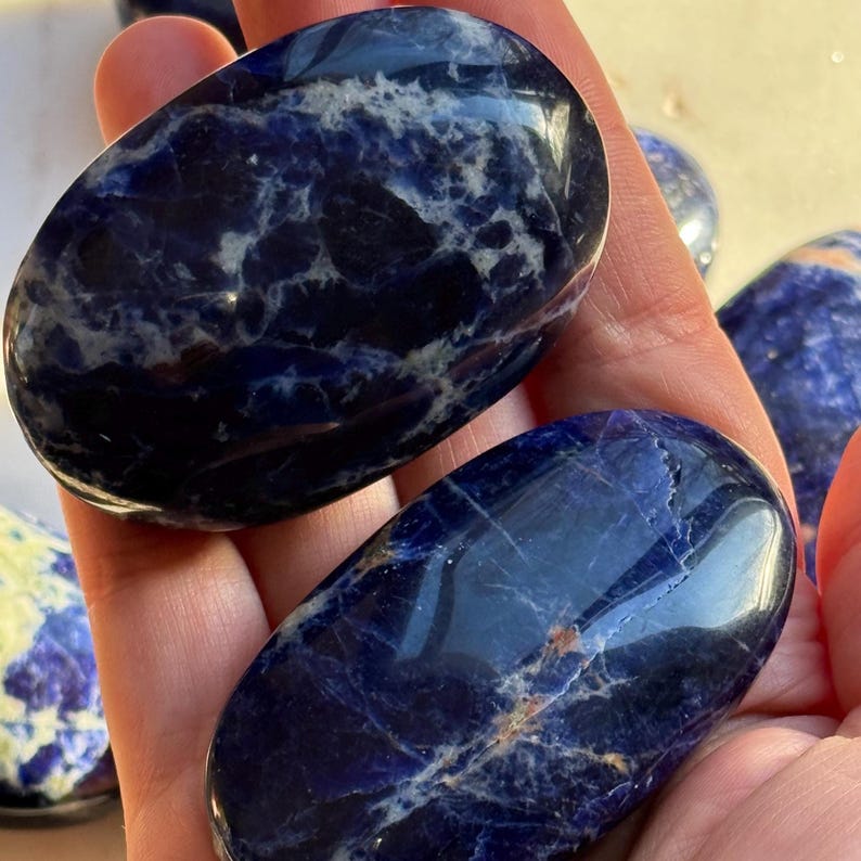 May include: Several polished sodalite stones in shades of deep blue and white are held in a hand. The stones have a smooth, oval shape and a marbled appearance. The natural stones are a popular choice for crystal healing and meditation.