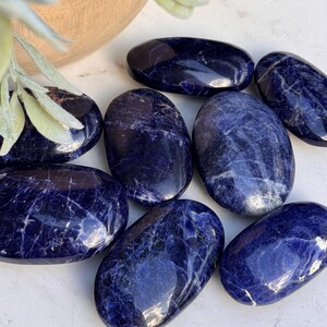 May include: A collection of polished sodalite stones. The stones are oval-shaped with a deep blue color and white veining. The stones are arranged on a white surface, with a wooden bowl and greenery in the background.