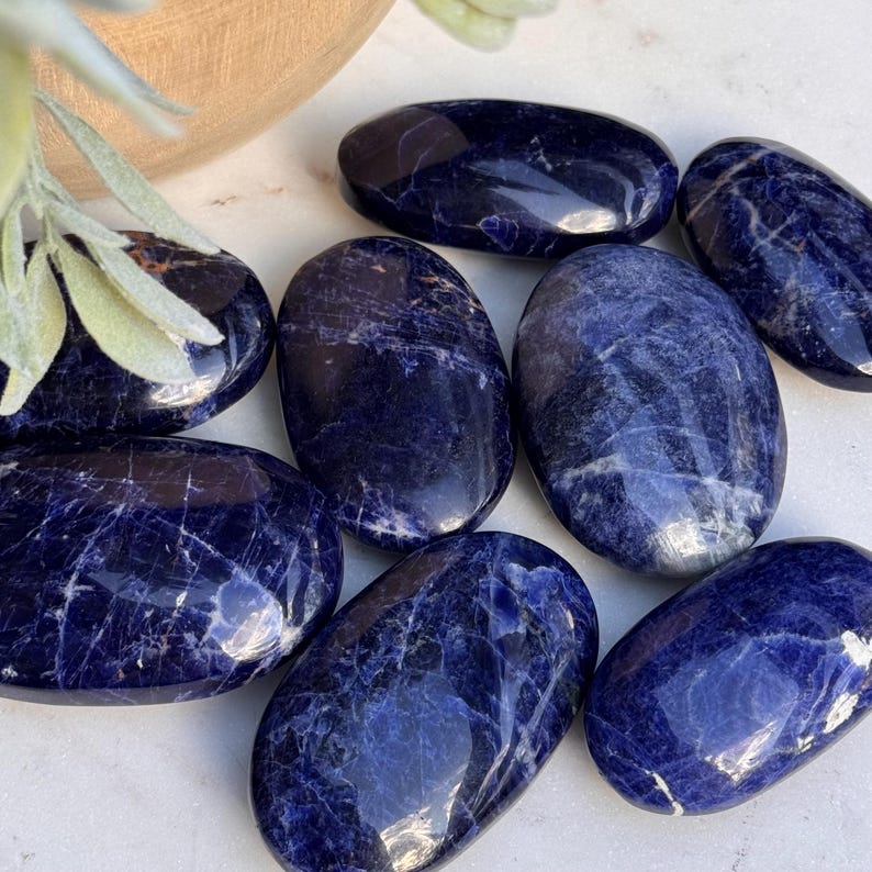 May include: A collection of polished sodalite stones. The stones are oval-shaped with a deep blue color and white veining. The stones are arranged on a white surface, with a wooden bowl and greenery in the background.