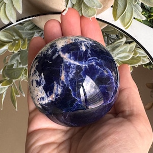 May include: A polished sodalite sphere, predominantly deep blue with white and tan veining. The sphere is held in a hand, reflecting the surrounding environment. The gemstone is a decorative item.
