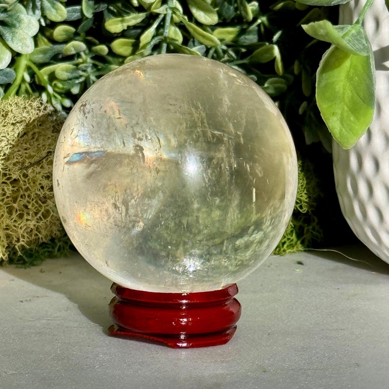 Op de afbeelding: Een heldere kristallen bol met interne breuken staat op een glanzende, rode houten standaard. De bol heeft een lichte gele tint. Groen gebladerte en een witte, getextureerde vaas zijn op de achtergrond.