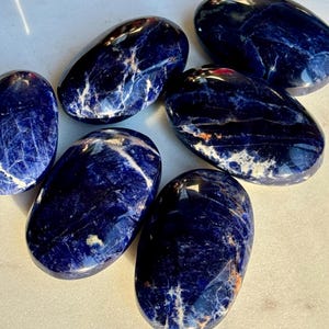 May include: A collection of polished sodalite stones. The stones are oval-shaped with a deep blue color, veined with white and hints of orange. The smooth, glossy surfaces reflect light, highlighting the natural patterns.