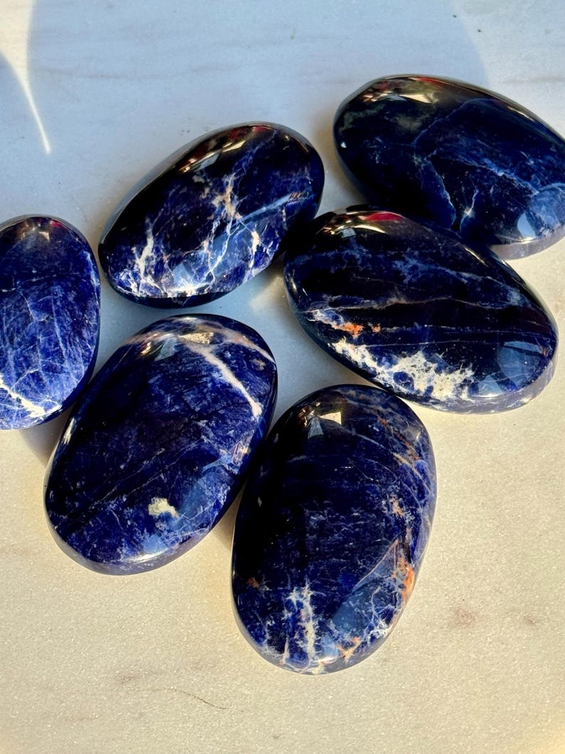 May include: A collection of polished sodalite stones. The stones are oval-shaped with a deep blue color, veined with white and hints of orange. The smooth, glossy surfaces reflect light, highlighting the natural patterns.