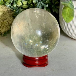 Op de afbeelding: Een heldere kristallen bol, met groene en witte insluitsels, rust op een glanzende, rode houten standaard. De achtergrond bevat groen gebladerte en een witte decoratieve pot.