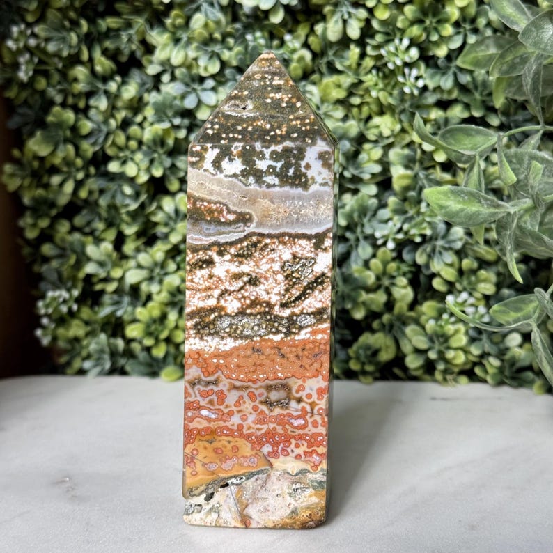May include: A rectangular, polished stone obelisk with a pointed top. The stone features bands of color including white, brown, orange, and green. The obelisk is standing upright against a backdrop of green foliage.