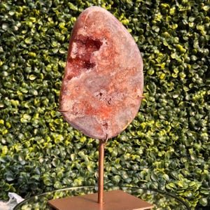 May include: A polished, irregular, reddish-orange geode slice with a central crystalline cavity, mounted on a thin, vertical copper-toned metal rod attached to a square base. The stone features varied textures, translucent edges, and deep red hues.