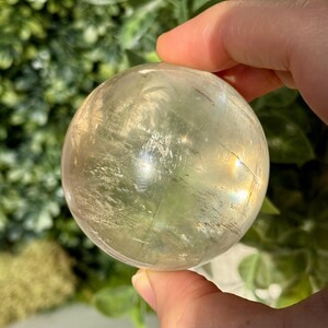 Op de afbeelding: Een heldere, bolvormige kristallen bol met een lichtgele tint wordt in een hand gehouden. Het kristal heeft interne breuken en insluitsels en reflecteert licht, waardoor een subtiel regenboogeffect ontstaat. De achtergrond is wazig en toont groen gebladerte.