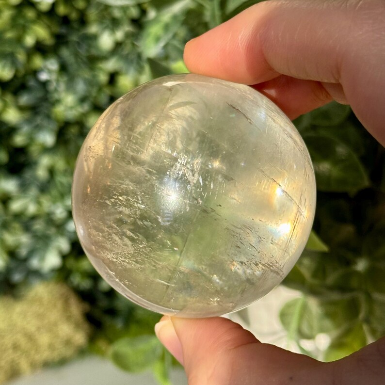 Op de afbeelding: Een heldere, bolvormige kristallen bol met een lichtgele tint wordt in een hand gehouden. Het kristal heeft interne breuken en insluitsels en reflecteert licht, waardoor een subtiel regenboogeffect ontstaat. De achtergrond is wazig en toont groen gebladerte.