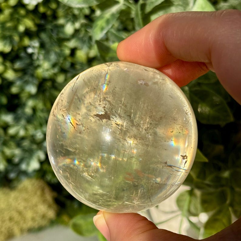 Op de afbeelding: Een heldere, bolvormige kristallen bol met een gele tint. De kristallen bol heeft interne insluitsels en vertoont regenboogbrekingen. De achtergrond is een vage mix van groen gebladerte. De kristallen bol wordt door een hand vastgehouden.