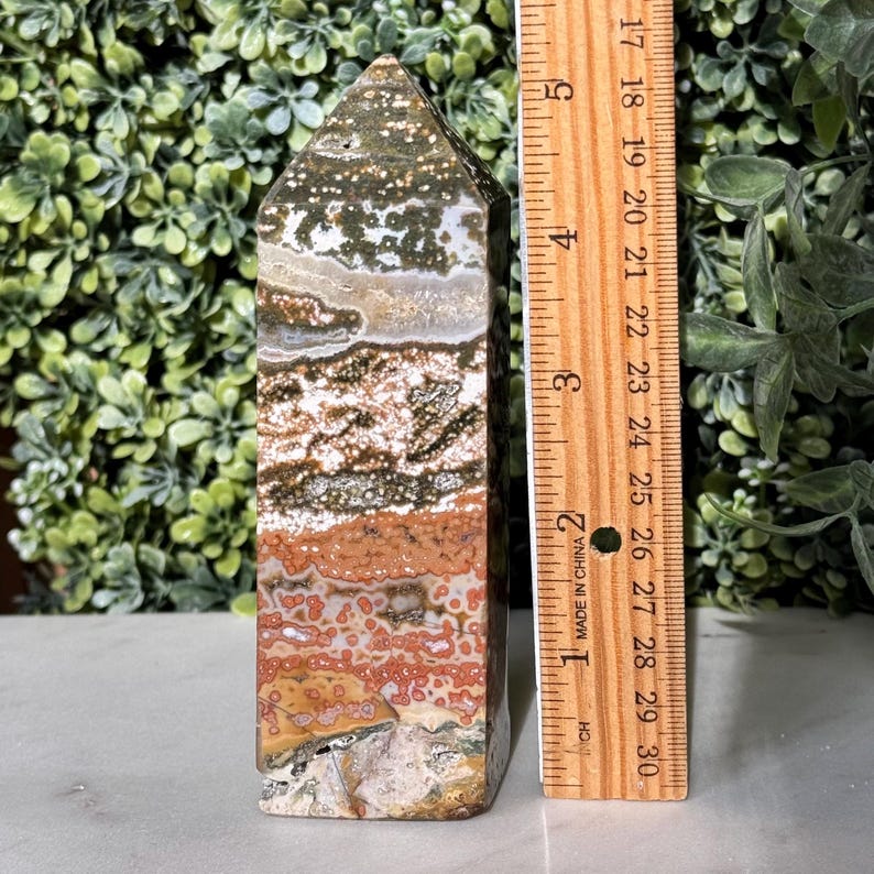 May include: A polished ocean jasper obelisk, approximately 5 inches tall, featuring a mix of earthy tones including brown, red, and green. The obelisk has a rectangular base and a pointed top, with a natural, layered pattern.