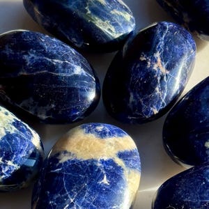 May include: A collection of polished sodalite stones. The stones are oval-shaped with a deep blue color and white and tan veining. The smooth, glossy surfaces reflect light, highlighting the natural patterns. These stones are often used for their aesthetic appeal and metaphysical properties.