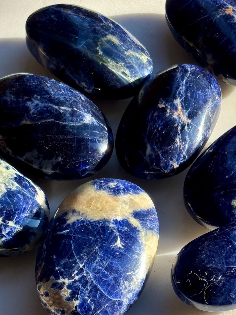 May include: A collection of polished sodalite stones. The stones are oval-shaped with a deep blue color and white and tan veining. The smooth, glossy surfaces reflect light, highlighting the natural patterns. These stones are often used for their aesthetic appeal and metaphysical properties.