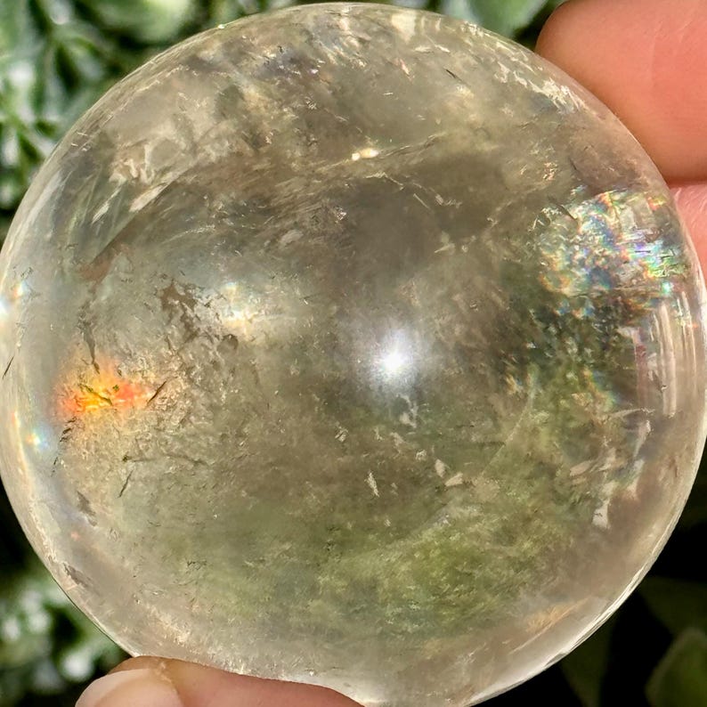 Op de afbeelding: Een heldere, bolvormige kristallen bol met interne insluitsels en regenboogirisatie. Het gepolijste oppervlak reflecteert licht, waardoor een glinsterend effect ontstaat. De kristal wordt tegen een achtergrond van groen gebladerte gehouden.